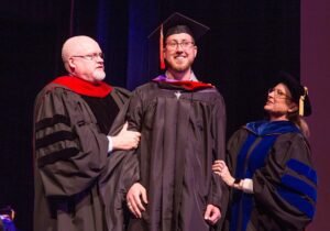 ĵֱ graduate student at graduation standing between two professors smiling.